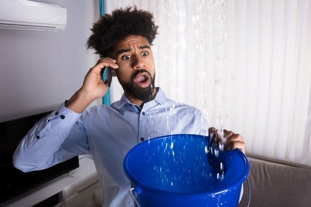 A man indoors, looking shocked, holds a phone to his ear and a blue bucket under a stream of water from the ceiling. He wears a light blue shirt.