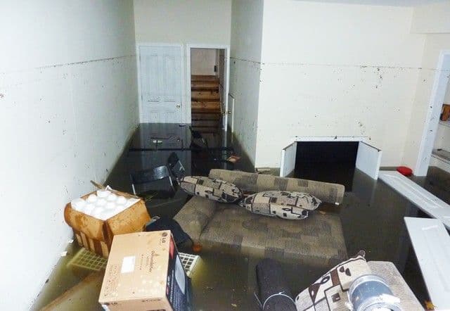 Flooded basement with floating furniture, a submerged sofa, and scattered boxes. Waterlogged walls show waterline, creating a chaotic, disordered scene.