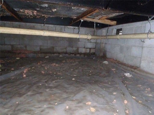 A dimly lit basement with concrete block walls and exposed piping overhead. The floor is covered with a plastic sheet and scattered debris, creating an abandoned feel.