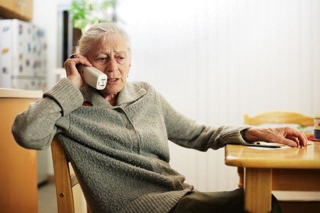 Elderly woman in a gray sweater sits at a wooden table, holding a phone to her ear, appearing contemplative. The kitchen in the background is softly lit.