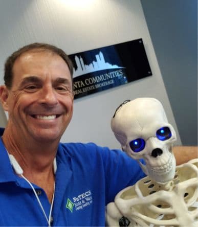 A man in a blue shirt smiles alongside a skeleton with glowing blue eyes. They stand in front of a sign for a real estate brokerage. Fun, quirky atmosphere.