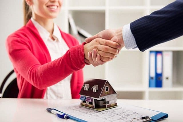 A woman in a red cardigan and a man in a suit shake hands over a desk. A small house model and floor plans are visible, suggesting a real estate deal.