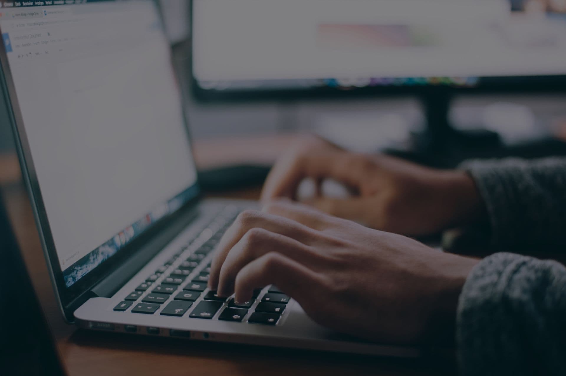 Hands typing on a laptop keyboard with a focused screen in the background. The setting suggests a workspace environment, conveying productivity.