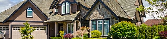 A charming suburban house with a stone facade and steep roof, surrounded by a lush garden with manicured bushes and vibrant greenery under a blue sky.