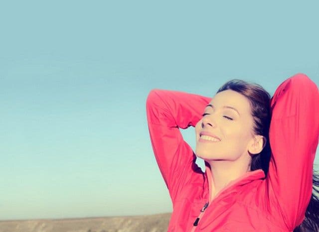 A woman in a red jacket stands outdoors against a clear blue sky, eyes closed and smiling, arms stretched behind her head, expressing relaxation and joy.