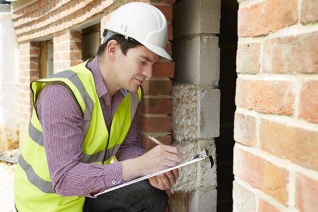 A construction inspector in a white hard hat and yellow safety vest examines a brick wall, focused and taking notes on a clipboard, conveying diligence.