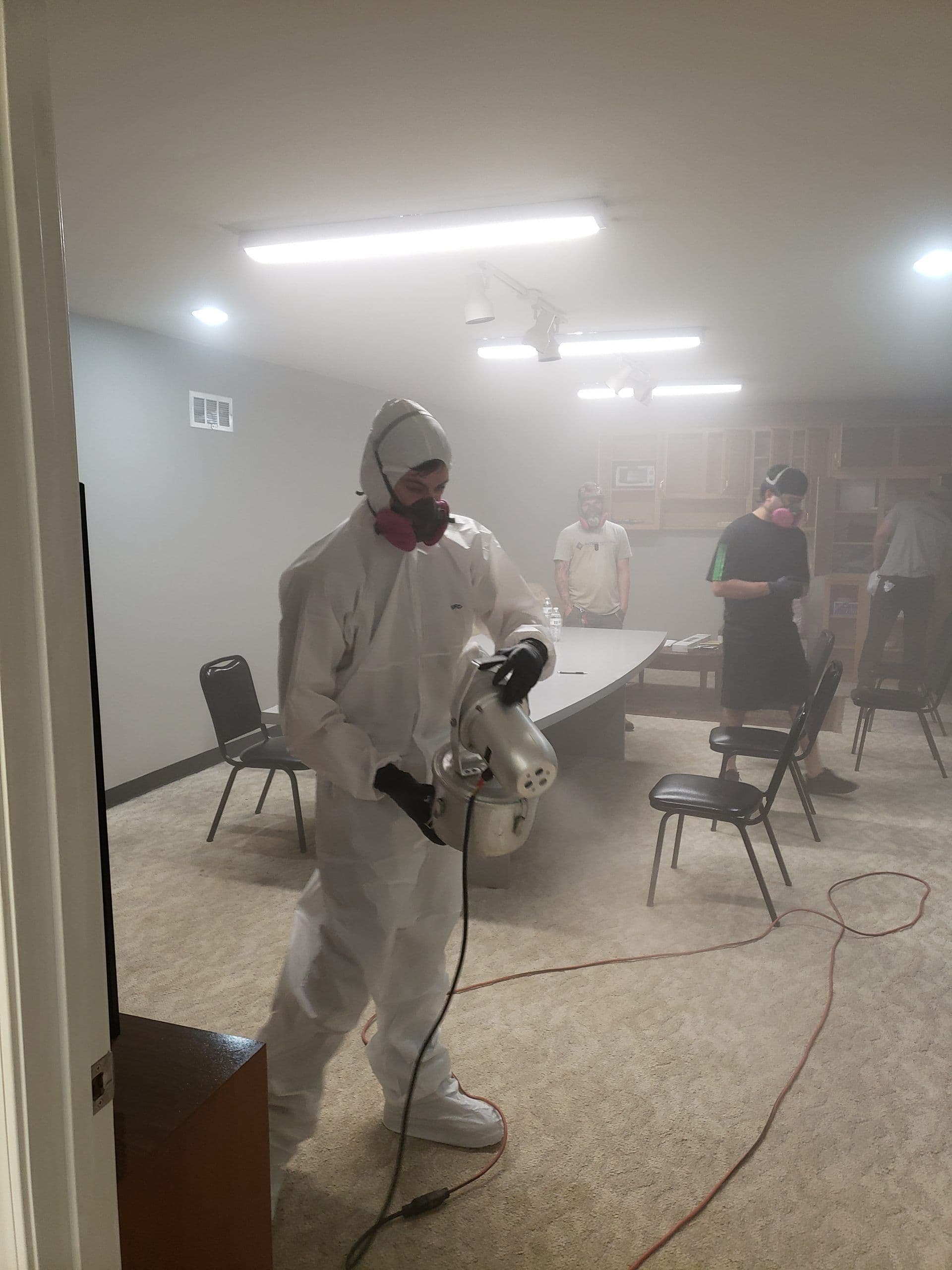 Person in white protective suit and mask fogging a room with a disinfectant machine; others stand nearby, room slightly hazy, serious atmosphere.