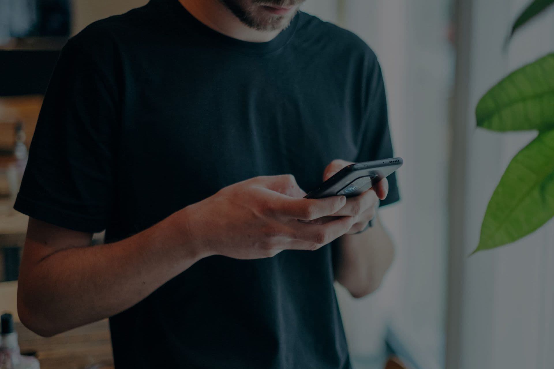 A person in a black T-shirt is focused on a smartphone, typing with both hands. They are indoors near a window with soft natural light.