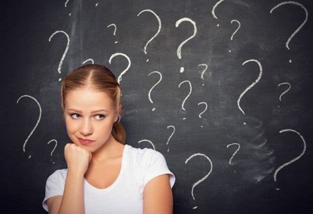 A woman in a white shirt is pondering, her hand on her chin, with a backdrop of numerous question marks on a blackboard, conveying curiosity and confusion.